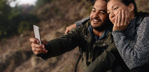 Horizontale Aufnahme eines verliebten jungen Paares beim Wandern, das ein Selbstporträt mit dem Smartphone im Freien macht. Entspanntes Wandererpaar im Urlaub, das ein Selfie mit dem Handy macht. - JLPSF28006