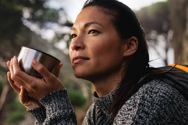 Nahaufnahme einer Wanderin, die sich mit einem Kaffee im Freien ausruht und wegschaut. Asiatische Frau beim Kaffee trinken. - JLPSF28011