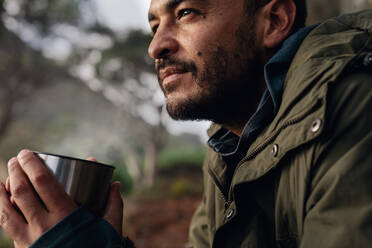 Nahaufnahme eines jungen kaukasischen Mannes mit Kaffee in der Hand, der beim Wandern eine Pause einlegt. Männlicher Wanderer, der sich im Freien ausruht. - JLPSF28012