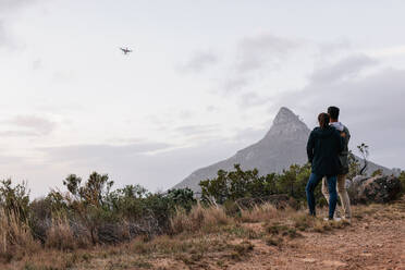 Junges Paar, das zusammen im Freien steht und eine fliegende Drohne am Himmel über der Landschaft beobachtet und steuert. Mann und Frau benutzen eine Drohne zum Fotografieren in ländlicher Umgebung. - JLPSF28036