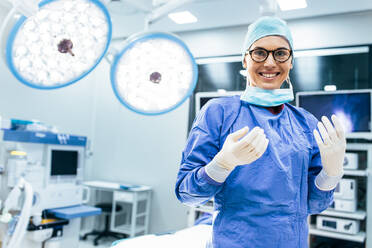 Portrait of happy woman surgeon standing in operating room, ready to work on a patient. Female medical worker in surgical uniform in operation theater. - JLPSF28043