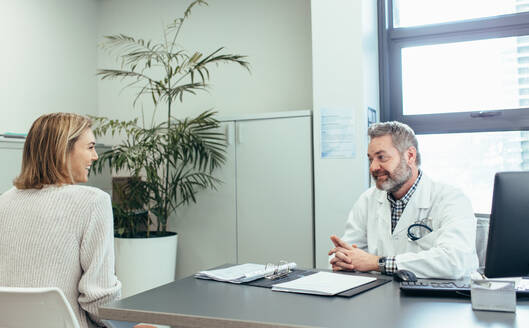 Glücklicher Arzt im Gespräch mit einer Frau, die am Schreibtisch sitzt. Freundlicher Arzt und Frau während der Konsultation in der Klinik. - JLPSF28047