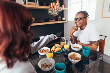 Glückliches junges Paar beim gemeinsamen Frühstück zu Hause: Frau gibt ihrem lächelnden Freund am Frühstückstisch ein Handy. - JLPSF28053