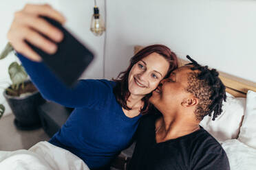 Romantisches multirassisches Paar liegt zusammen auf dem Bett und macht ein Selfie. Lächelnder Mann küsst auf die Wange der Frau, die ein Selfie mit dem Handy macht. - JLPSF28058