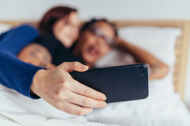 Paar, das zusammen auf dem Bett liegt und ein Selfie mit dem Smartphone macht. Fokus auf das Handy in der Hand der Frau. - JLPSF28059