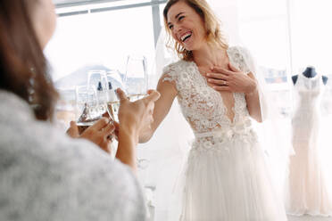 Fröhliche junge Frau im Hochzeitskleid stößt mit Freunden in einer Brautboutique mit Champagner an. Schöne Braut im eleganten Hochzeitskleid stößt mit ihren Freunden mit Champagner an und lächelt in einem Brautmodengeschäft. - JLPSF28067