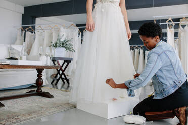 Female wedding dress designer making adjustment to bridal gown in her studio. Bride standing in her wedding gown with woman dressmaker making adjustments to gown. - JLPSF28074