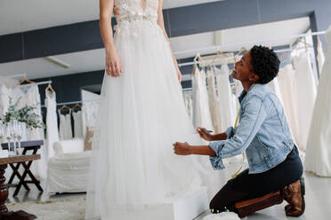 Woman making adjustment to bridal gown in wedding fashion store. Bride in her wedding dress with female dress designer making final adjustments on dress and smiling. - JLPSF28075