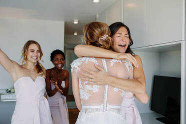 Glückliche Brautjungfern gratulieren der Braut im Hochzeitskleid im Hotelzimmer. Brautjungfern umarmen die Braut und lächeln. - JLPSF28085