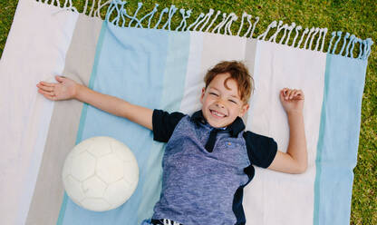 Fröhlicher Junge auf dem Boden liegend mit einem Fußball an seiner Seite. Draufsicht auf einen Jungen, der auf einer Decke in einem Park liegt. - JLPSF28093