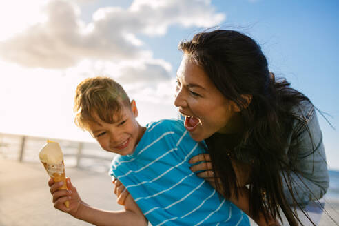 Junge isst mit seiner Mutter ein Eis in der Nähe der Strandpromenade. Kleiner Junge hält eine Eiswaffel, während seine Mutter spielerisch versucht, sie von hinten zu essen. - JLPSF28095