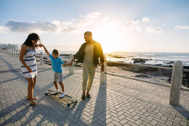 Fröhliche Familie im Urlaub, die den Sonnenuntergang im Freien genießt. Ein Paar hilft seinem Kind, am Meer Skateboardfahren zu lernen, indem es seine Hände hält. - JLPSF28097