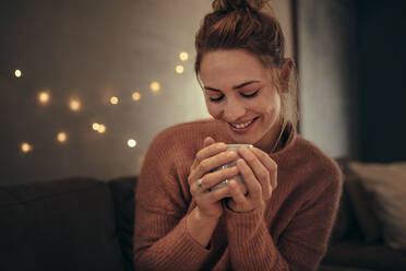 Lächelnde Frau trinkt Kaffee im Winter zu Hause. Kaukasische Frau entspannt sich im gemütlichen Wohnzimmer und trinkt Kaffee. - JLPSF28101