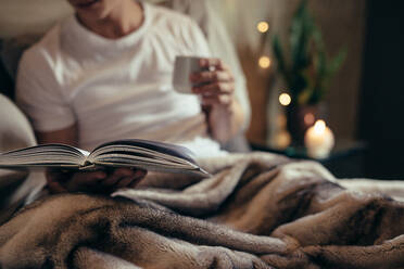 Nahaufnahme eines Mannes auf dem Bett mit offenem Buch und Kaffee. Ein Mann liest ein Buch auf dem Bett zu Hause. - JLPSF28103