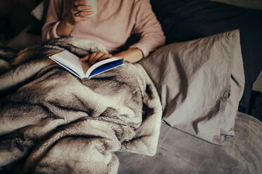Cropped shot of female on bed reading book and drinking coffee. Female reading book on bed at home. - JLPSF28104