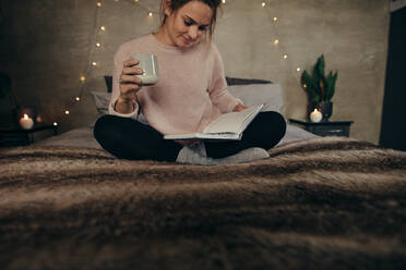 Entspannte junge Frau sitzt auf dem Bett, liest ein Buch und trinkt Kaffee. Frau liest ein Buch auf dem Bett zu Hause. - JLPSF28106