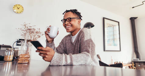 Happy young african man sitting at table using mobile phone and drinking coffee. Man relaxing at home reading text message on his cell phone in morning. - JLPSF28110