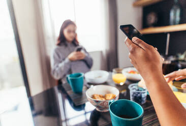 Nahaufnahme eines Mannes, der ein Mobiltelefon in der Hand hält, während er mit seiner Freundin im Hintergrund frühstückt, wobei der Fokus auf den Händen liegt, die das Smartphone halten. - JLPSF28117