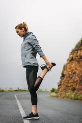 Fitness-Frau macht Dehnungsübungen auf einer leeren Straße. Frau im Sweatshirt streckt ihr Bein. - JLPSF28123
