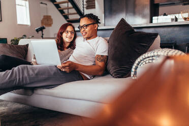 Mixed race couple sitting on a couch with laptop in living room. Couple relaxing on sofa at home using laptop computer. - JLPSF28124