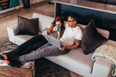 Cozy couple sitting on sofa at home. Woman drinking coffee and man with laptop while sitting on couch in living room. - JLPSF28127