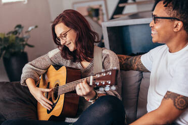 Hübsche junge Frau spielt Gitarre, während sie mit ihrem Freund auf dem Sofa sitzt. Paar entspannt zusammen zu Hause im Wohnzimmer. - JLPSF28136