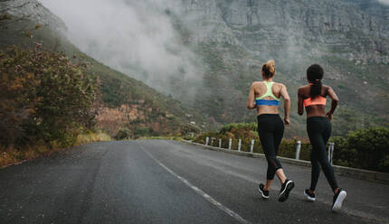 Zwei Frauen, die an einem nebligen Morgen auf der Straße laufen, mit Hügeln im Hintergrund. Fitness-Frauen joggen auf der Straße. - JLPSF28137