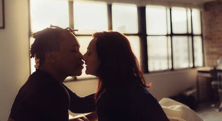 Zärtliches Paar, das sich im Schlafzimmer küsst. Interracial Mann und Frau sitzen auf dem Bett und küssen sich. - JLPSF28146