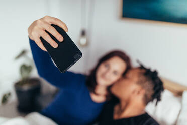 Paar, das zusammen auf dem Bett liegt und ein Selfie mit dem Handy macht. Fokus auf das Smartphone in der Hand der Frau. - JLPSF28147