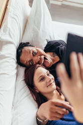 Top view of multiracial couple lying together on bed and taking selfie. Man sticking out tongue with woman taking selfie using mobile phone. - JLPSF28148