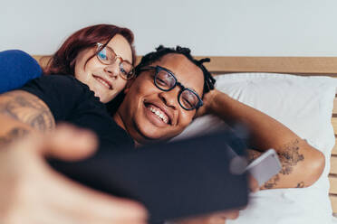 Fröhliches multirassisches Paar, das zusammen im Bett liegt und ein Selfie macht. Lächelnder Mann und lächelnde Frau machen ein Selfie mit Handy. - JLPSF28152