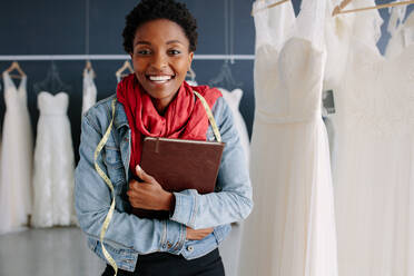 Fröhliche Brautladenbesitzerin in ihrer Boutique mit einem Tagebuch. Weibliche Hochzeitsmodedesignerin in einer Brautmodenboutique. - JLPSF28182