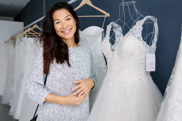 Attraktive junge Braut lächelnd bei der Auswahl Hochzeitskleid in Braut Boutique. Lächelnde asiatische Frau Einkaufen für Hochzeit Outfit in Braut Kleidung zu speichern. - JLPSF28183