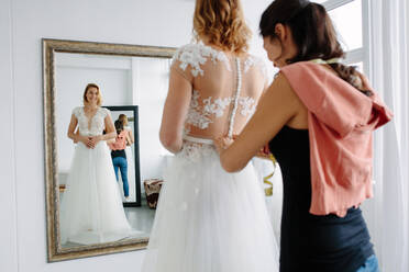 Spiegelbild einer Frau, die in einem Geschäft ein Hochzeitskleid anprobiert, mit einer Assistentin, die der Braut hilft, sich in ein Hochzeitskleid zu kleiden. - JLPSF28202