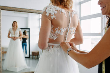 Die Braut steht vor einem Spiegel und passt ihr Kleid in einem Atelier mit einem Hochzeitsassistenten an. Eine Frau mit einem Schneider macht den letzten Schliff an einem maßgeschneiderten Kleid in einem Brautmodengeschäft. - JLPSF28204