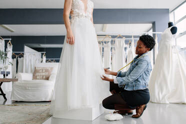 Afrikanische Frau, die Anpassungen am Hochzeitskleid im Atelier eines Modedesigners vornimmt. Die Braut steht in ihrem Hochzeitskleid mit einer Designerin, die letzte Anpassungen am Kleid vornimmt. - JLPSF28215