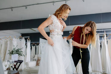 Female making adjustment to wedding gown in fashion designer studio. Bride wearing her wedding gown with female dress designer making final adjustments on dress. - JLPSF28219