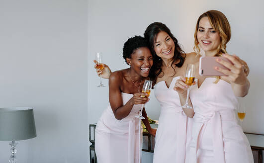 Drei Brautjungfern, die ein Selfie in einem Raum machen. Lächelnde junge Frauen, die etwas trinken und ein Selfie mit dem Handy machen. - JLPSF28238