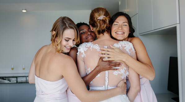 Fröhliche Brautjungfern umarmen Braut im Hochzeitskleid im Hotelzimmer. Hochzeitstag, glückliche Brautjungfern gratulieren der Braut und lächeln. - JLPSF28241