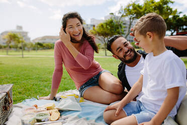 Ehepaar mit Kind beim Picknick; Mann und Frau mit Kind genießen Snacks im Freien in einem Park. - JLPSF28251