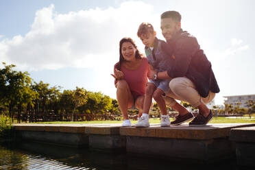 Familie in fröhlicher Stimmung in einem Park. Paar mit ihrem Kind sitzt am Ufer eines Teiches und schaut auf das Wasser. - JLPSF28255