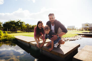 Familie in fröhlicher Stimmung in einem Park. Paar mit Kind sitzt auf der Brücke eines Baches und schaut auf das Wasser. - JLPSF28256