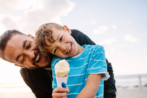 Junge isst mit seinem Vater ein Eis in der Nähe der Strandpromenade. Kleiner Junge hält eine Eistüte, während sein Vater spielerisch versucht, sie von hinten zu essen. - JLPSF28262