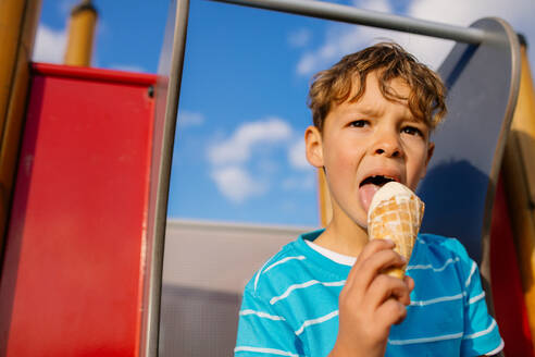 Nahaufnahme eines Jungen, der auf einer Rutsche sitzend ein Eis isst. Kleiner Junge im Urlaub, der sich ein Eis gönnt. - JLPSF28265