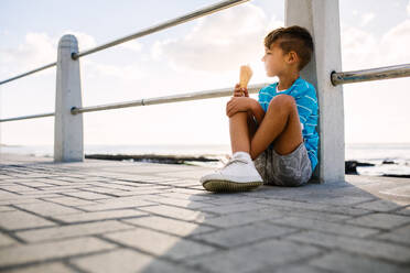 Junge isst ein Eis an einem Geländer am Meer mit der untergehenden Sonne im Hintergrund. Kleiner Junge im Urlaub, der sich ein Eis gönnt. - JLPSF28267