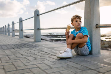 Junge isst ein Eis, sitzt an einem Geländer am Meer und schaut weg. Kleiner Junge im Urlaub, der sich ein Eis gönnt. - JLPSF28268