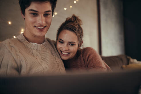 Lächelnder junger Mann und Frau mit Laptop zu Hause. Gemütliches Paar auf dem Sofa sitzend mit einem Laptop im gemütlichen Wohnzimmer. - JLPSF28279