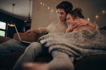 Junges Paar, das im Winter zu Hause auf dem Sofa sitzt und einen Laptop benutzt. Gemütliches Paar in einem hyggeligen Haus mit Laptop. - JLPSF28280