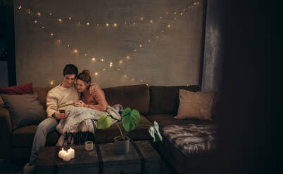 Verliebtes Paar sitzt auf dem Sofa und benutzt das Handy zu Hause. Mann und Frau entspannen sich auf der Couch mit Smartphone im gemütlichen Wohnzimmer. - JLPSF28281