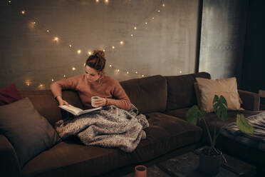 Young woman reading book and drinking coffee on sofa in hygge house. Caucasian female relaxing in cozy living room and reading a book. - JLPSF28282